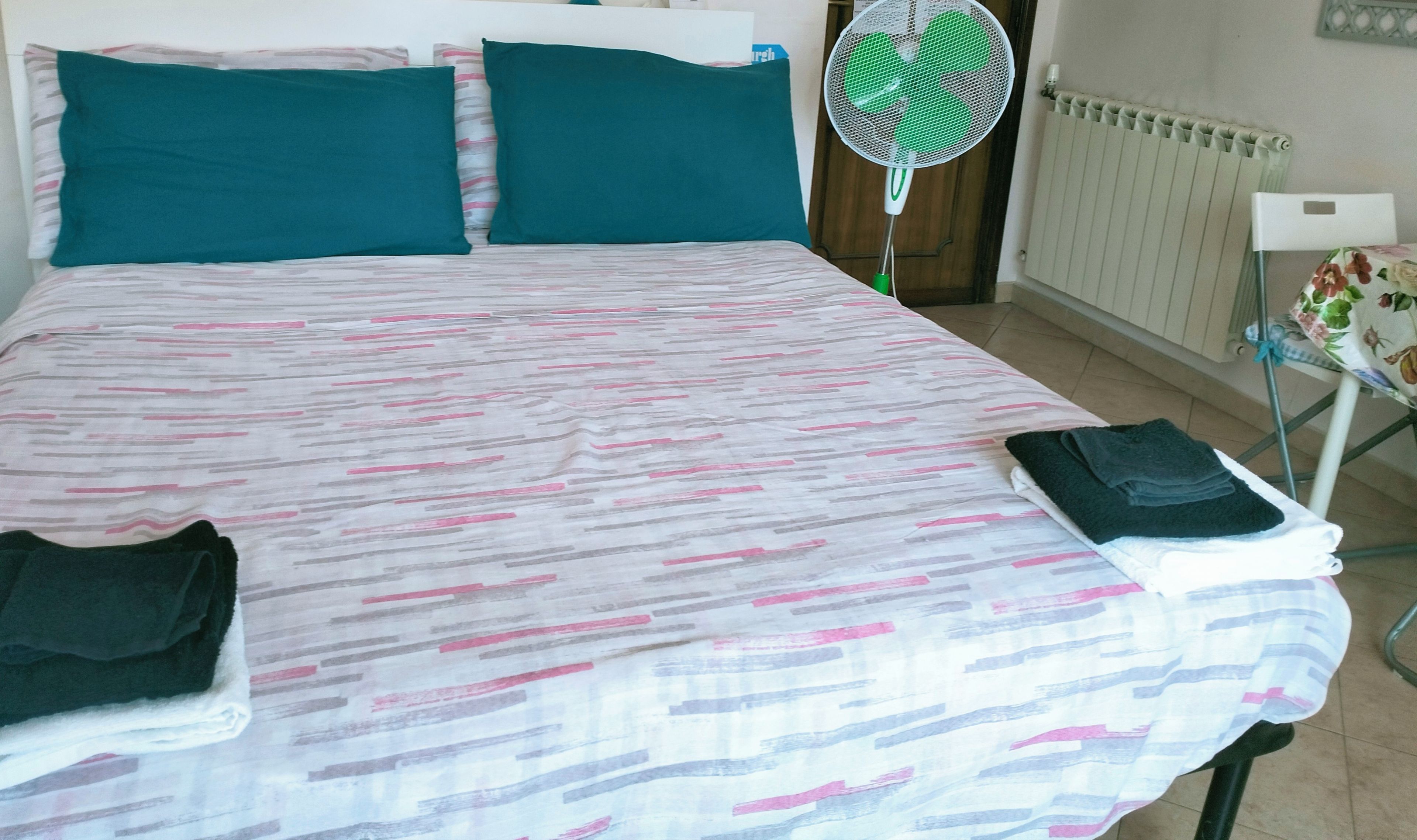 Habitación de hotel con cama, almohadas turquesas, cartel con la hora de registro y un ventilador de pie, además de un radiador.