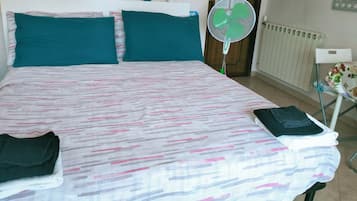 Habitación de hotel con cama, almohadas turquesas, cartel con la hora de registro y un ventilador de pie, además de un radiador.