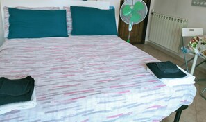 A hotel room with a bed, teal pillows, a check-in time sign, a standing fan, and a radiator. - La Perla B & B (Ciampino)