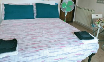Habitación de hotel con cama, almohadas turquesas, cartel con la hora de registro y un ventilador de pie, además de un radiador.