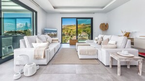 A modern living room with two white sofas, a coffee table, and a TV. Large windows offer a view of a coastal landscape. - Marbella Holiday Villa with Sea Views & Heated Pool, Gym (AN)