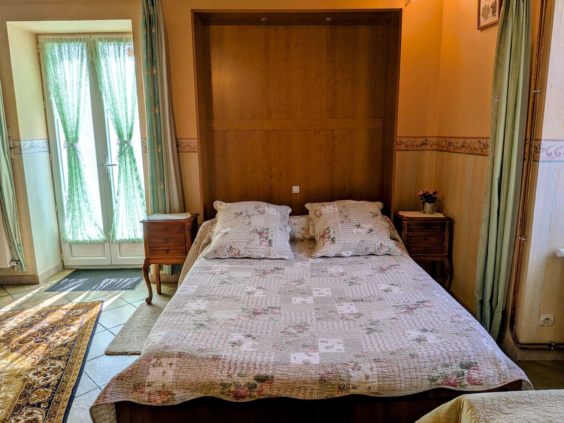 Une chambre à coucher avec un grand lit, deux tables de chevet, une tête de lit en bois, une fenêtre avec des rideaux et un tapis à motifs.