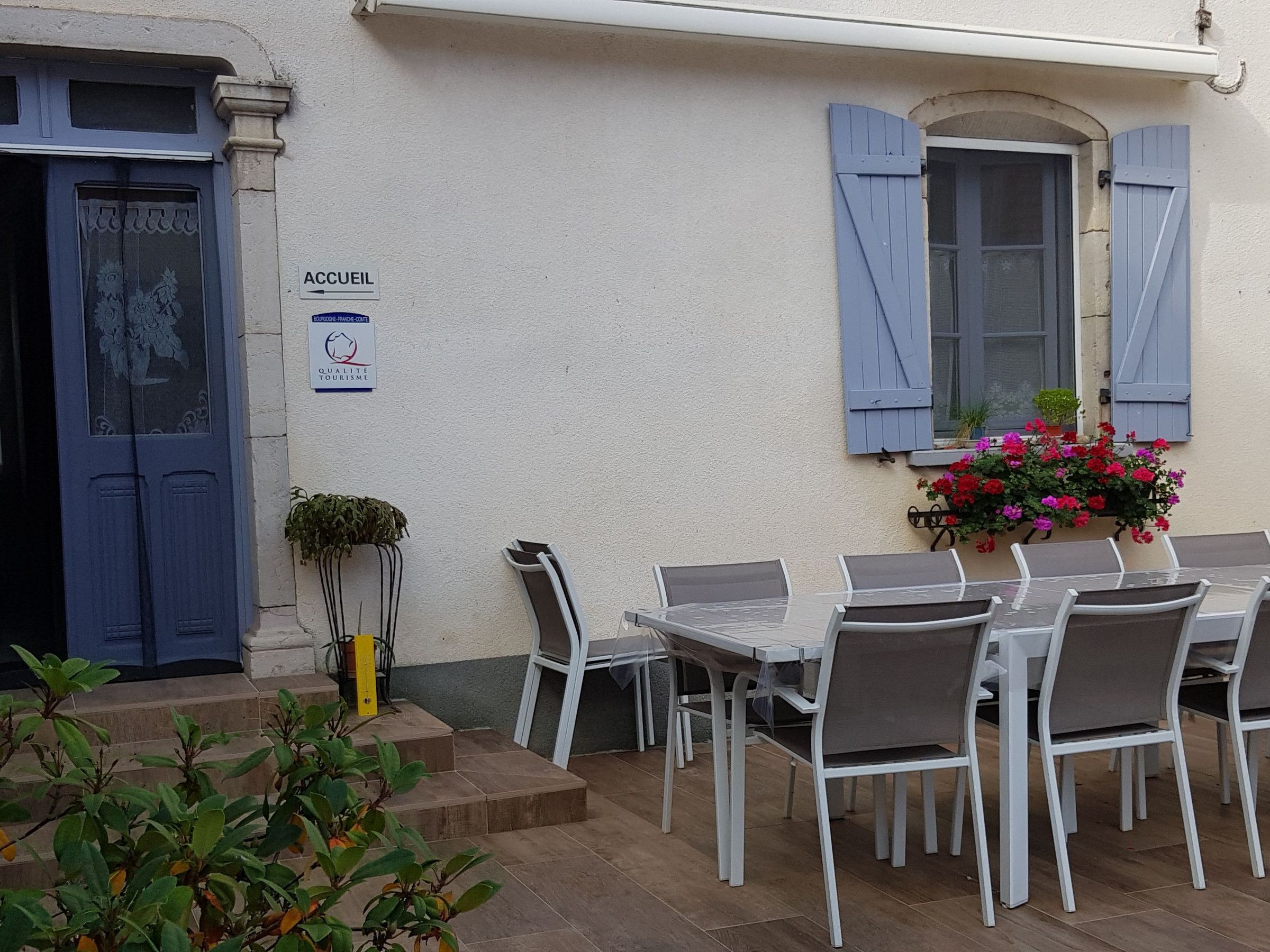 Un espace extérieur aménagé avec des tables et des chaises blanches, une porte bleue et une enseigne indiquant « ACCUEIL ».