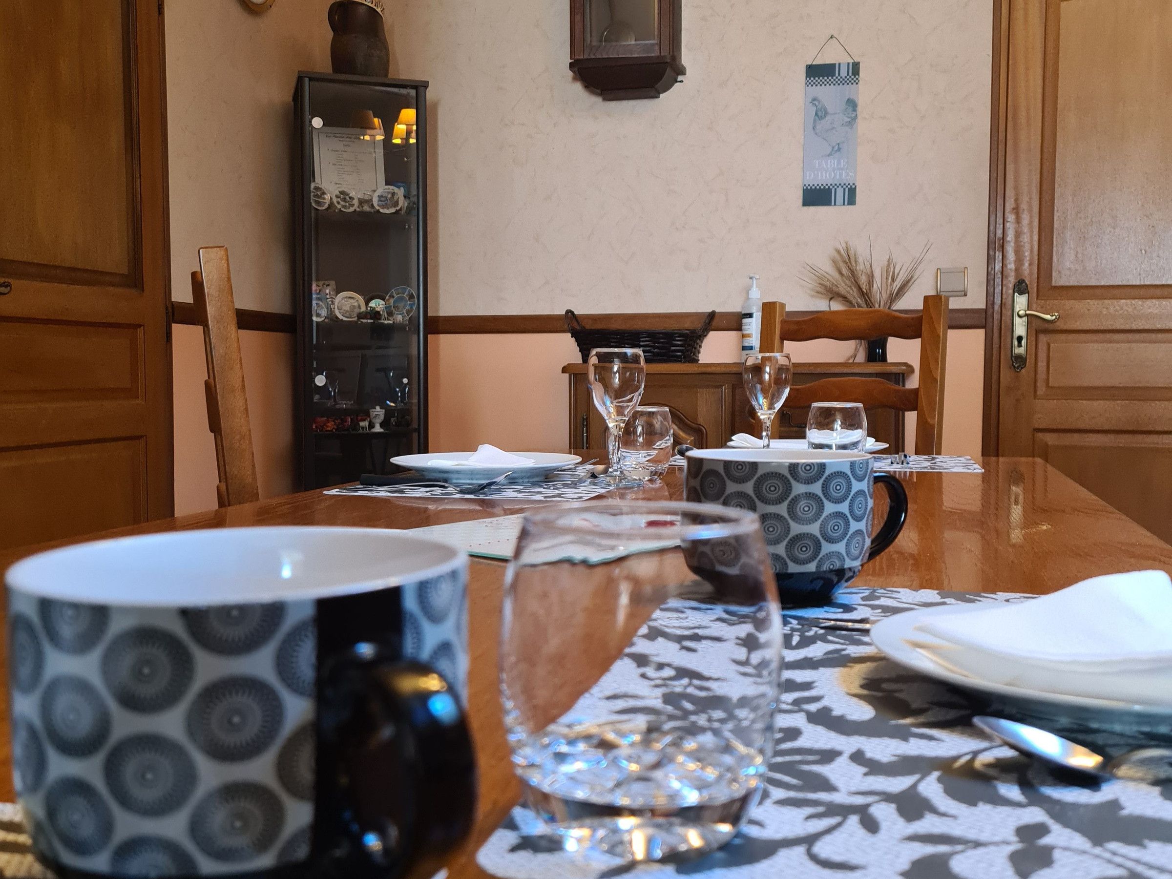 Een eettafel gedekt met borden, glazen en een karaf. Er is een kast met servies en een bordje aan de muur.