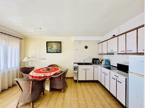 A compact kitchen with white cabinets, a dining area with wicker chairs, a red tablecloth, and a painting on the wall.