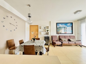 A dining area with a table and chairs, a sofa, a wall clock, a shelf with decorative items, and a painting.