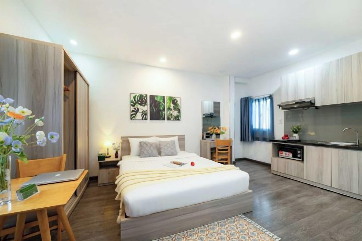 A modern hotel room with a large bed, a desk with a laptop, a small dining table, and a kitchenette.