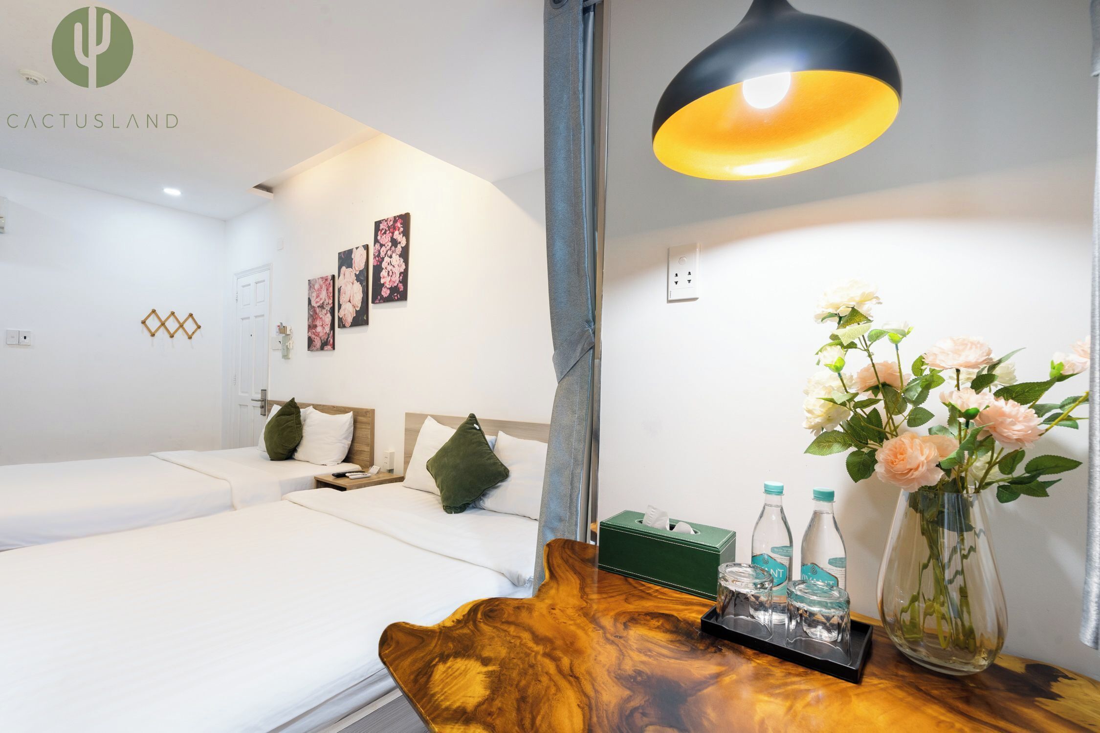 Ein Hotelzimmer mit zwei Betten, einem Holfnachtkästchen mit Blumen in einer Vase, einer grünen Papiertütenbox und einer schwarzen Lampe.