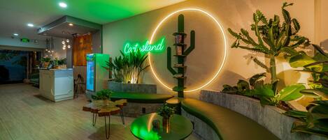 Un hall d’entrée avec une enseigne au néon indiquant « Cartland », un comptoir d’accueil, un réfrigérateur et diverses plantes.