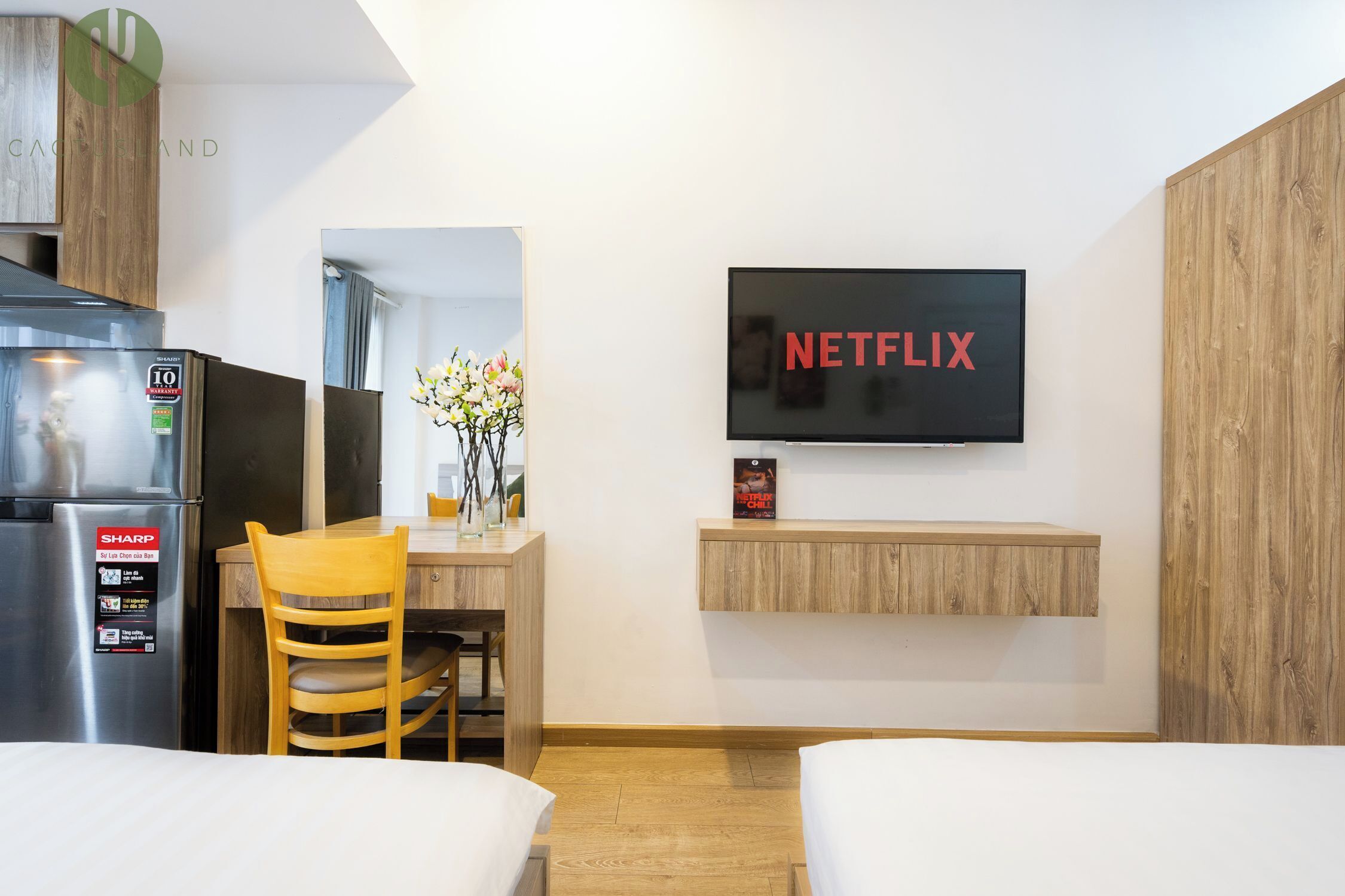 Habitación de hotel con cama, escritorio con silla, refrigerador, televisor montado en la pared y estante de madera.