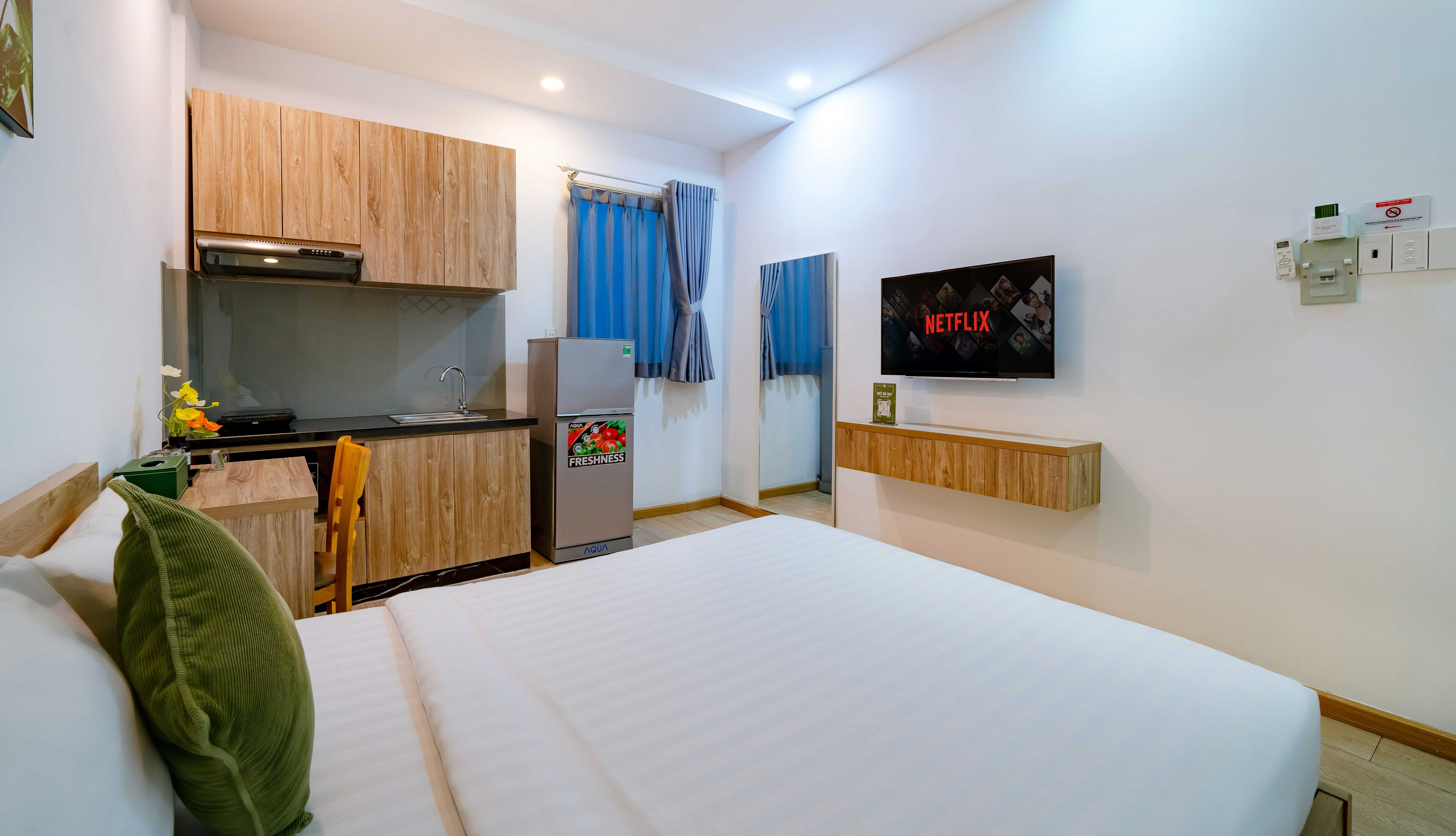 A compact hotel room with a kitchenette, a bed, a television, and a wall-mounted control panel.