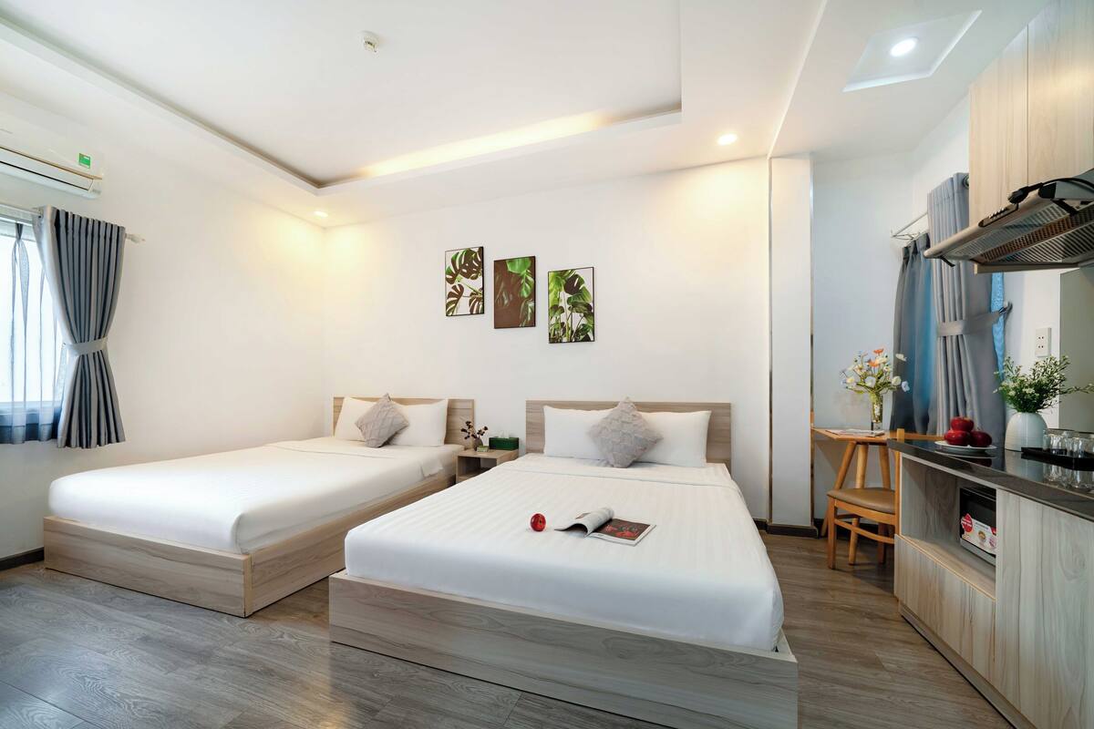 Une chambre d’hôtel avec deux lits simples, une petite table avec une chaise, un coin cuisine et des œuvres d’art accrochées aux murs.
