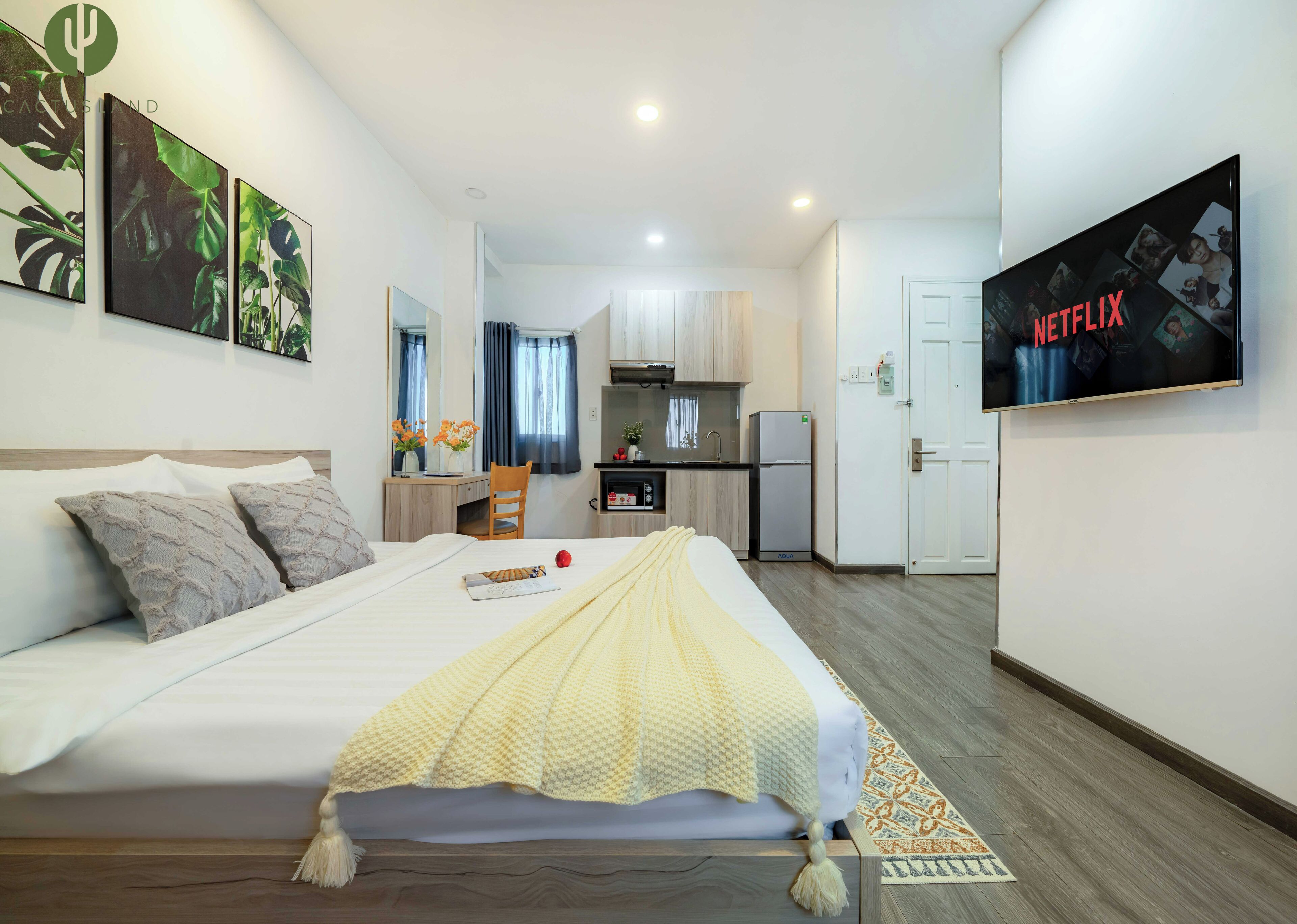 Quarto de hotel com cama, escrivaninha, cozinha compacta e televisão exibindo 'NETFLIX'.
