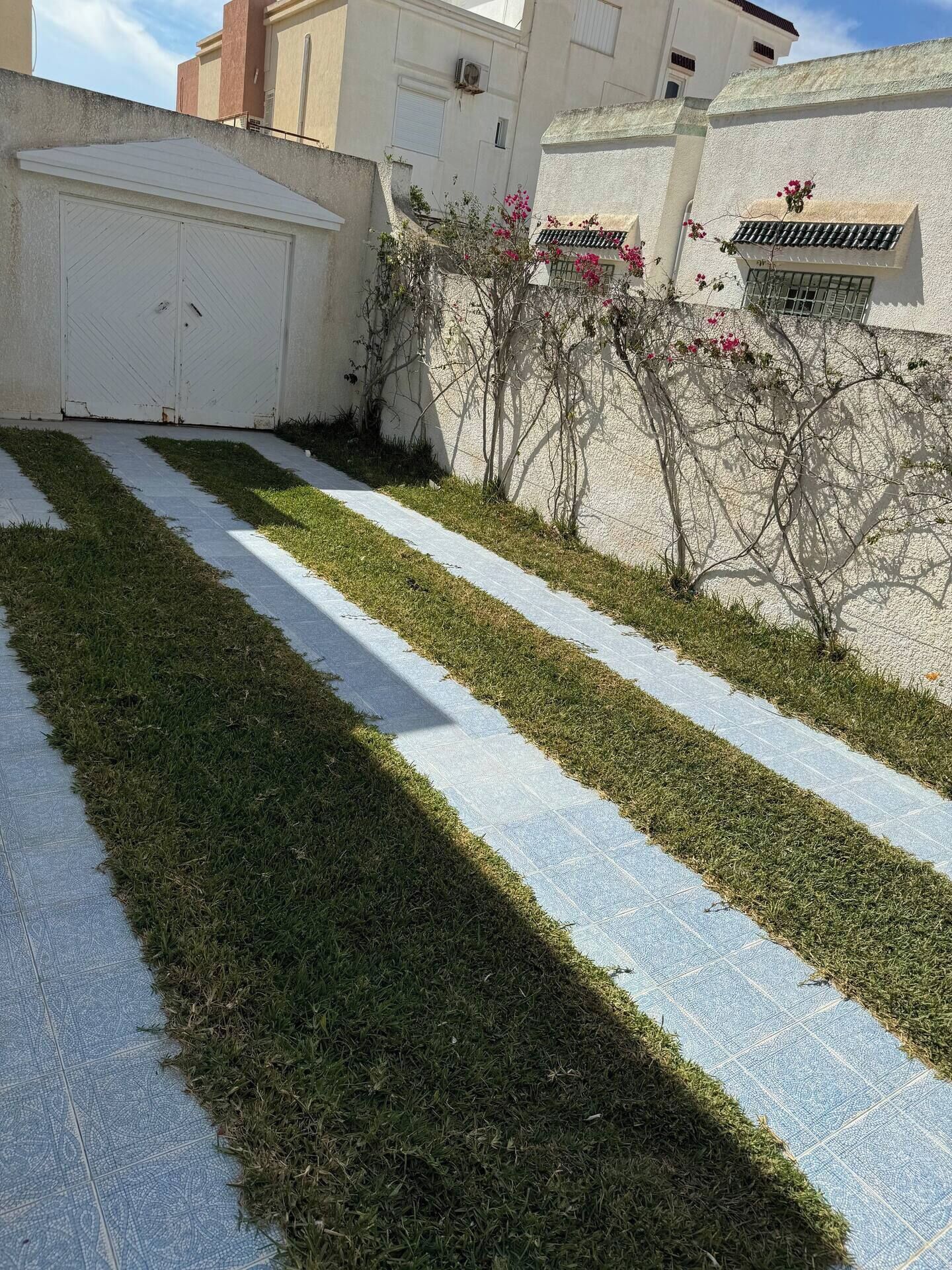 A paved pathway with alternating sections of grass and concrete, bordered by a low wall with a white garage door and a building with a tiled roof.