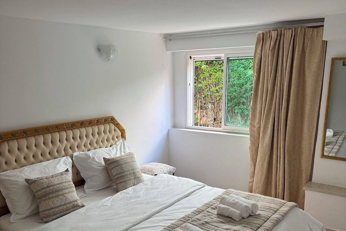 Una cama bien hecha con una colcha y almohadas estampadas, una ventana con cortinas y un espejo en la pared.