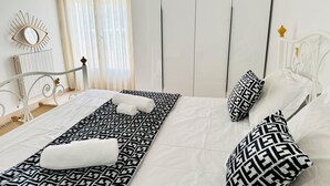 A neatly made bed with patterned bedding, a decorative mirror, and a window with sheer curtains.