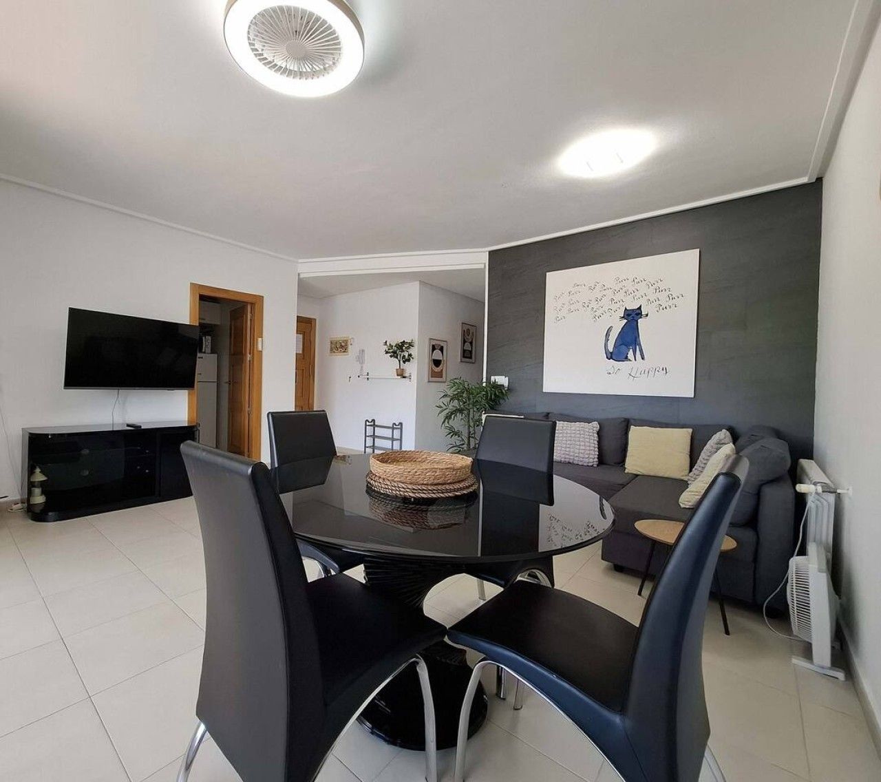 A modern living room with a dining table, chairs, a sofa, a flat-screen TV, and a wall-mounted artwork.