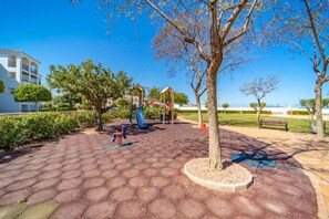 A playground with a slide and swings, surrounded by trees and a paved area. - Murcia Vacations`s Apartments 1, Apartamento fa... (La Torre Golf Resort)