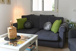 Una sala de estar con un sofá gris oscuro, cojines verdes, una mesa de centro de vidrio con revistas y un farol de madera.