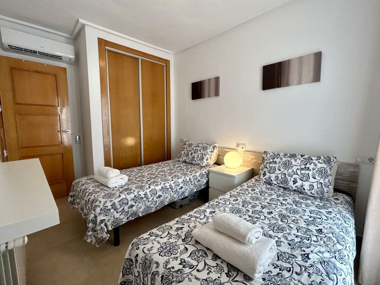 Une chambre avec deux lits, une armoire, une table de chevet avec une lampe et un luminaire fixé au mur.