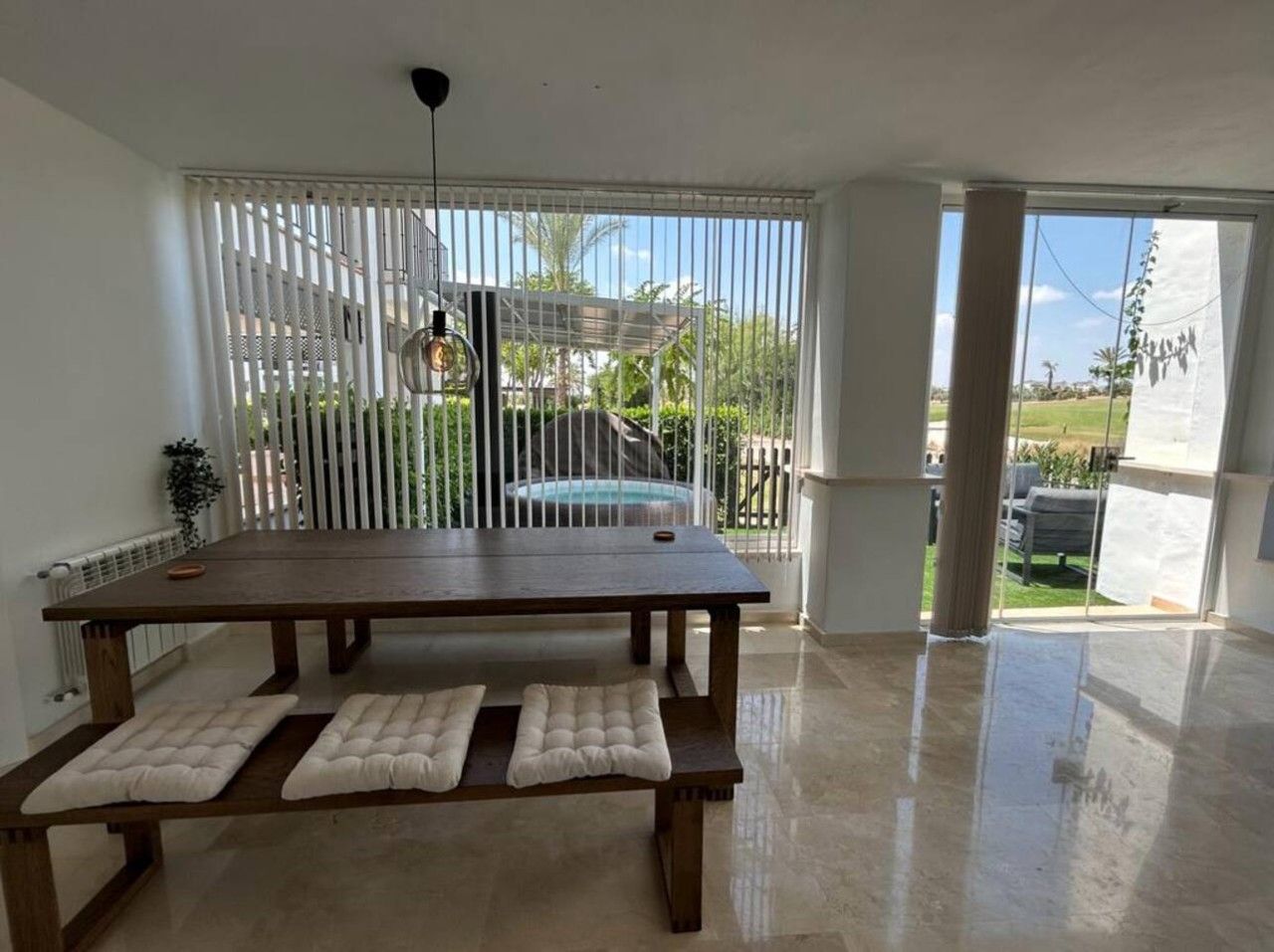 A spacious room with a large wooden table, cushioned benches, and a view of a backyard through a sliding glass door.