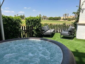 A hot tub with two lounge chairs, a wooden fence, and a view of a golf course.
