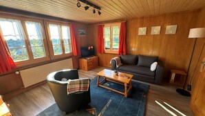 A cozy living room with a sofa, a coffee table, and a TV. There are windows with curtains, a radiator, and a lamp. - Ferienwohnung Gehrihorn by Interhome (Frutigen)