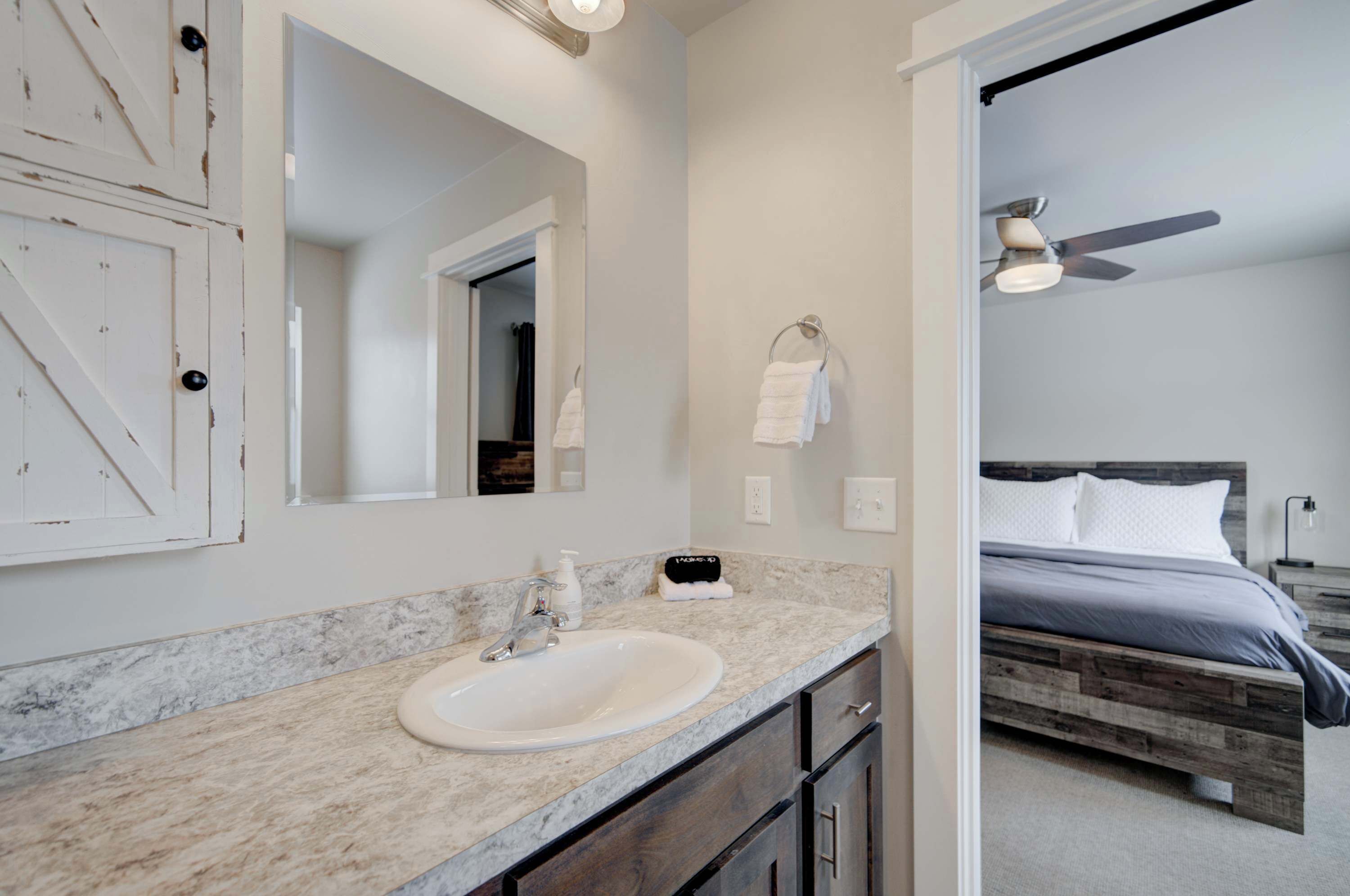 A bathroom with a marble countertop, a white sink, and a mirror. There is a wooden bed with a headboard in the adjacent room.