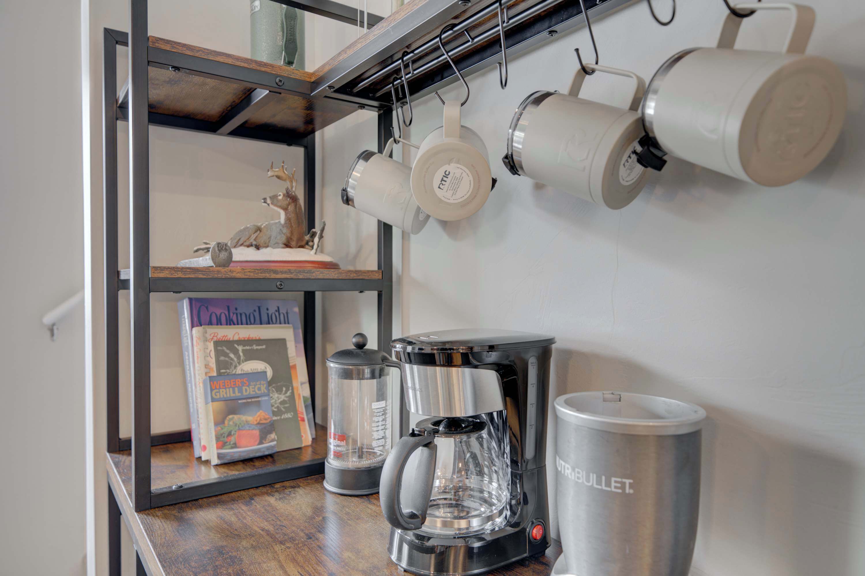 A kitchen area with a coffee maker, a kettle, and hanging pots. There is a book titled "Weber's Grill Deck" on a shelf.