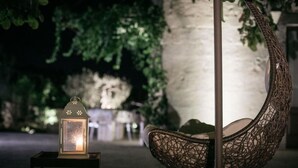 Une chaise suspendue avec une structure en osier, une lanterne posée sur une petite table et un mur de pierre en arrière-plan.