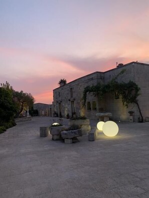 Une cour extérieure avec un bâtiment en pierre, de grandes lampes sphériques et un ciel au coucher du soleil.
