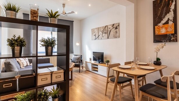 A modern living room with a sofa, TV, dining table, and chairs. There are potted plants on shelves and a ceiling fan. - Nice apartment in Vila de Gracia (Barcelona)