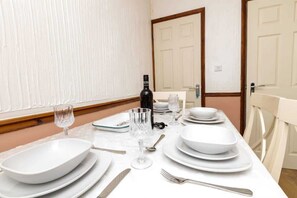 Une table à manger dressée avec de l’assiette blanche, des bols, des couverts et des verres à vin, ainsi qu’une bouteille de vin et un verre posés sur la table.