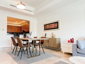 A modern dining area with a wooden table and chairs, a kitchen in the background, and a framed artwork on the wall.