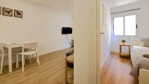 Una habitación con zona de comedor que incluye una mesa y sillas blancas, un televisor de pantalla plana montado en la pared y un ventanal que proporciona luz natural.