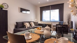 A modern living room with a glass dining table, dark sofas, and a large window.