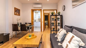 A modern living room with a wooden coffee table, a grey sofa with decorative pillows, a bookshelf with potted plants, and a dining area with chairs and a table.