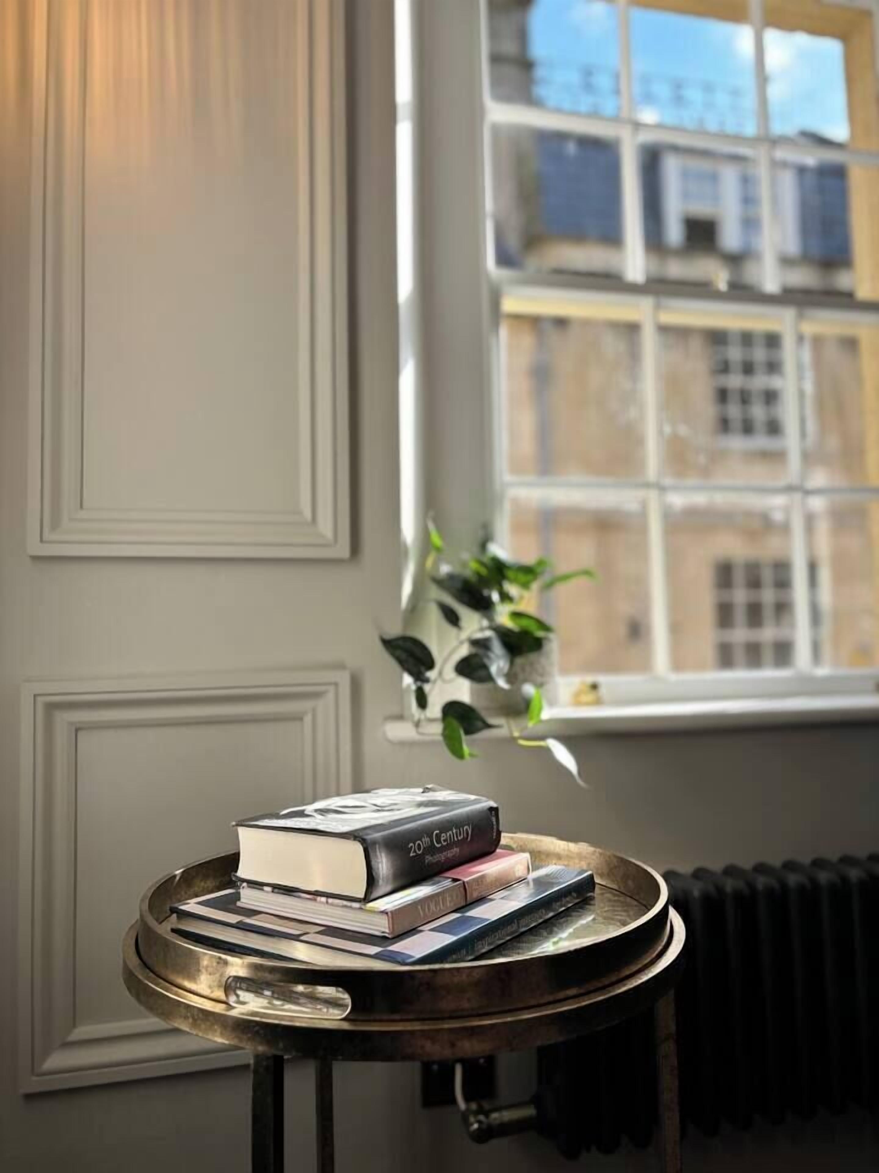 A round table with a stack of books and a potted plant in a room with a window and radiator.