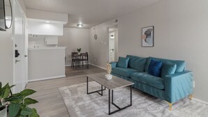 Un salon moderne avec un canapé de couleur turquoise, une table basse en bois et une cuisine en arrière-plan.