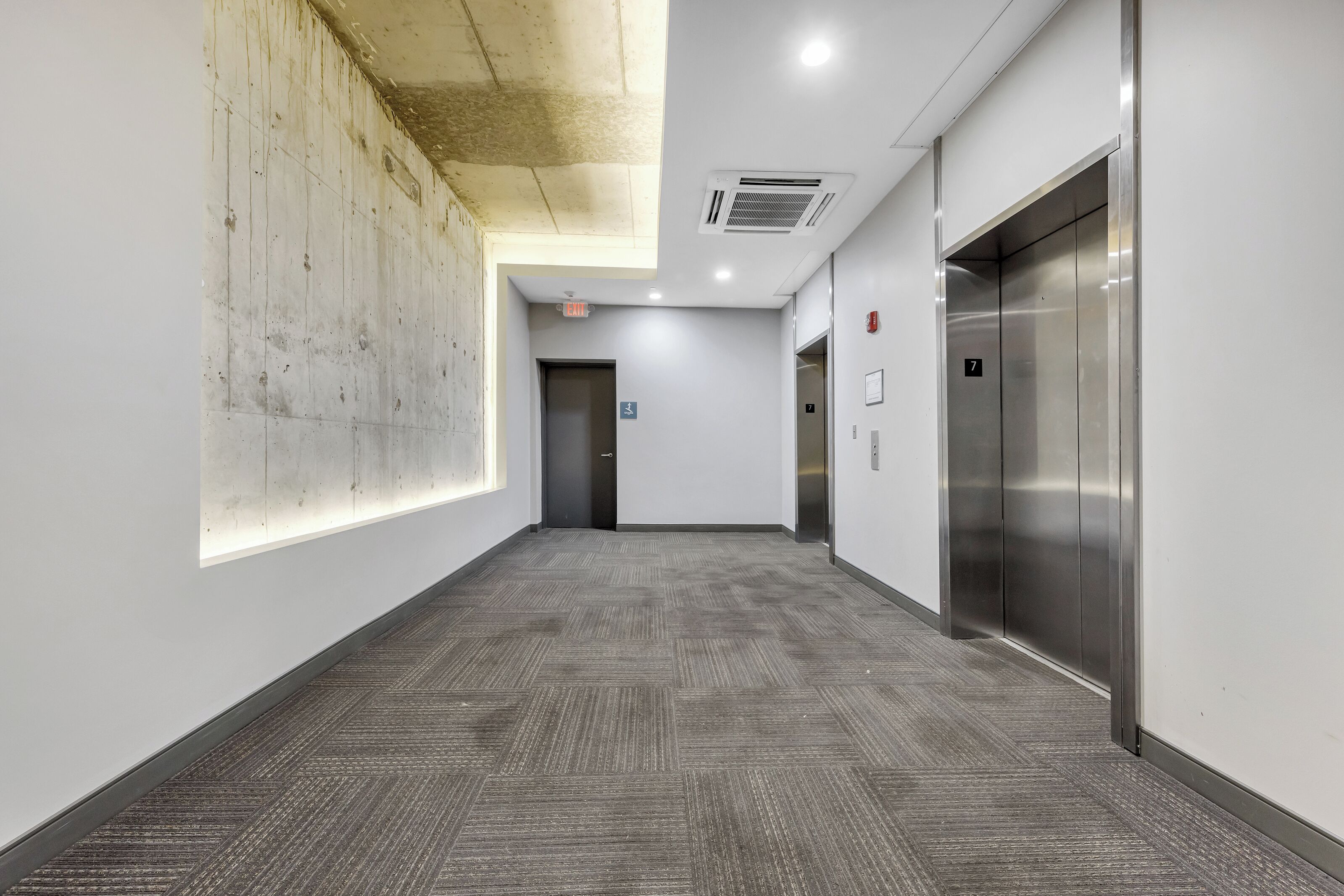 A hallway with a carpeted floor, a wall with a textured pattern, and an elevator on the right.