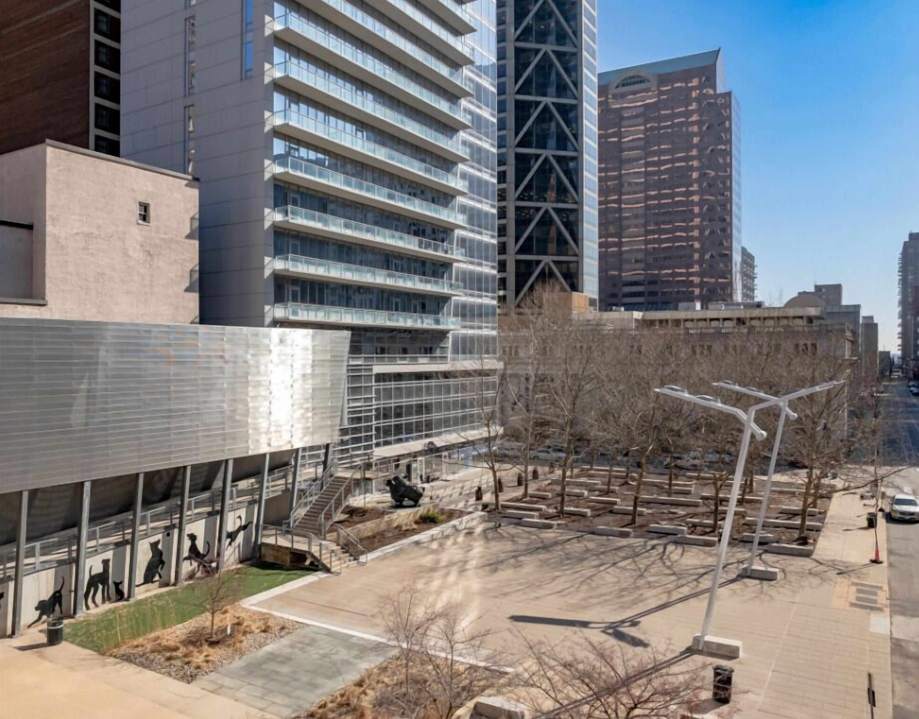 A modern urban plaza with a large glass building, a metal structure with animal sculptures, and a clear sky.