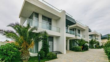 Un moderno edificio residencial de varios pisos con balcones, entrada pavimentada y abundante vegetación.