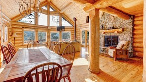 A spacious wooden cabin with a large dining table, multiple chairs, a stone fireplace, and large windows offering views of the surrounding forest.