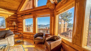 A log cabin interior with a sofa, two leather armchairs, a lamp, and large windows offering mountain views.