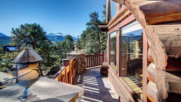 A wooden cabin with a stone pathway leading to a glass door, surrounded by mountains and trees.
