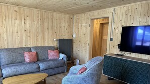 Een kamer met houten lambrisering, een grijze bank, een blauwe fauteuil, een houten salontafel en een televisie op een kast.