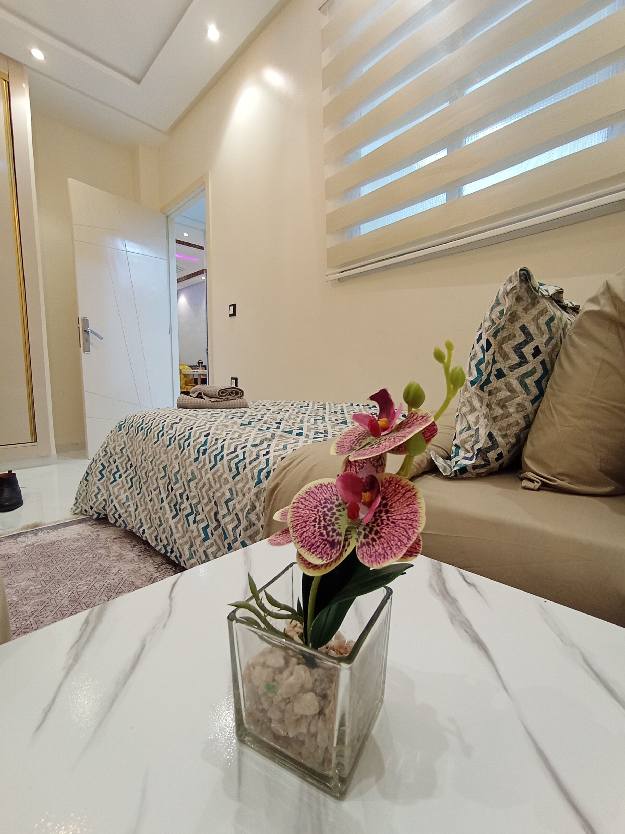 A modern hotel room with a bed, a sofa, a glass vase with flowers, and a window with blinds.