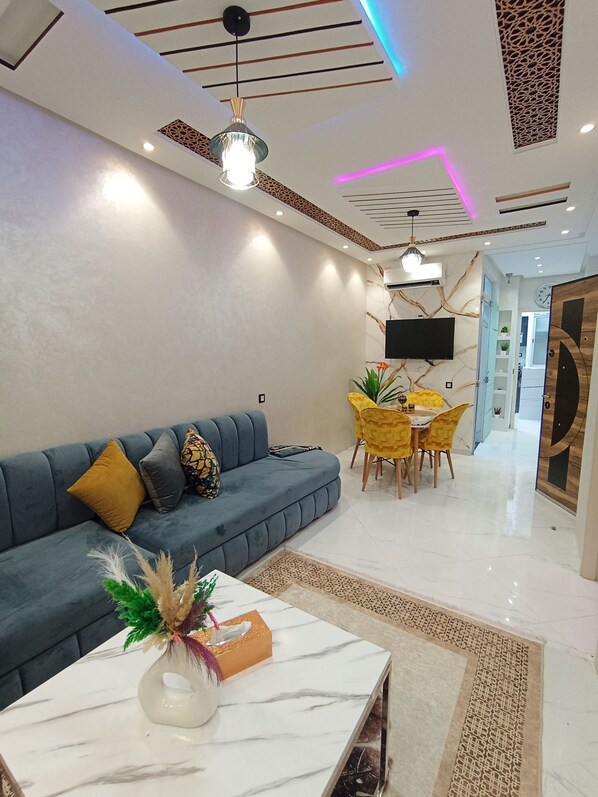 A modern living room with a blue sofa, a white coffee table, and a dining area with yellow chairs. - Aphrodite Apartment (Tangier)