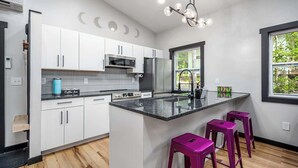A modern kitchen with white cabinets, a central island with bar stools, a built-in oven, and a window with a view of greenery. - Two Unique Treehouses in Nature (Pittsboro)