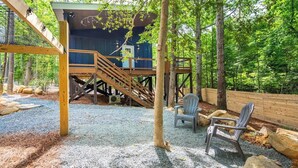 A wooden deck with a staircase leading to a blue building, surrounded by trees and outdoor seating. - Two Unique Treehouses in Nature (Pittsboro)